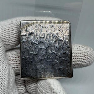 May include: A silver-coloured, rectangular lighter with a textured, wave-like pattern on its surface. The lighter has a rectangular shape and appears to be made of metal. The design is detailed and the lighter is held by a gloved hand.