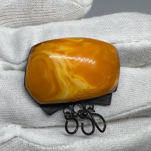 Op de afbeelding: Een broche met een grote, gepolijste, goudgeel en wit gemarmerde barnsteensteen op een donkere, geoxideerde metalen basis. De onderkant van de metalen zetting is voorzien van drie kleine, bungelende, cirkelvormige draadlussen.