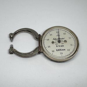 Peut inclure: Un pied à coulisse à cadran argenté avec une jauge circulaire et un bras de mesure incurvé. La jauge a des marquages noirs sur fond blanc, avec le texte "0-5 mm" et "0.025 mm".
