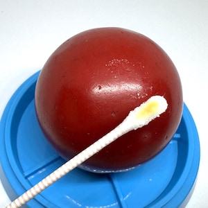 May include: A smooth, spherical red object rests on a circular blue plastic base. A white cotton swab with a yellow residue on its tip is positioned diagonally across the red sphere, showing a contrast between the red surface and the swab.