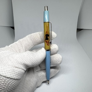 May include: A blue and yellow pen with a silver clip. The pen has a light blue top and bottom, with a yellow centre featuring a design. The pen is held by a gloved hand.