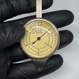 May include: A vintage map measurer with a silver handle and a circular dial. The dial has a cream-coloured face with black markings and a needle pointing to a distance in miles. The tool is held by a gloved hand.
