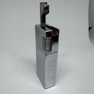 Może przedstawiać: Srebrna zapalniczka ZYKLOS METALBAU KG. Zapalniczka ma prostokątny korpus z teksturowanym wałkiem do zapłonu i otwieraną górą. Tekst "ZYKLOS METALBAU KG" jest wygrawerowany z boku.