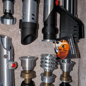 Puede incluir: Colección de réplicas de sables de luz plateados, negros y bronce. Las empuñaduras presentan diversos diseños, incluyendo formas acanaladas, cilíndricas y angulares. Algunos tienen botones rojos y secciones de emisor detalladas. También se ve una tarjeta de visita con una ilustración de un perro.