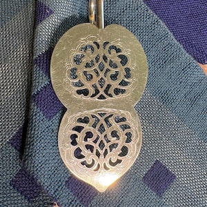 May include: A silver-toned metal cake server featuring a dual-lobed, flat head with intricate, symmetrical cut-out scrollwork patterns. The handle attachment is visible at the top, and the surface displays a polished, reflective metallic finish.