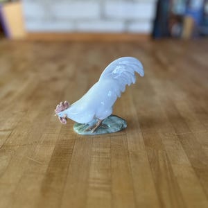 May include: A porcelain figurine of a white rooster with a light blue tint, featuring a detailed red comb and wattle, standing on a small, textured green base. The rooster is depicted in a forward-leaning posture with its tail feathers arched.