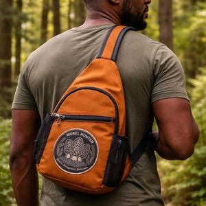Peut inclure: Un sac bandoulière orange avec une bordure noire et des poches latérales en filet. Le sac comporte un écusson avec le texte "Morel Hunter" et des illustrations de champignons. Le sac est porté sur l'épaule.