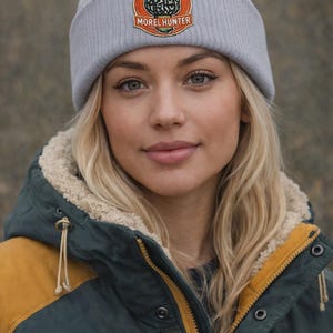 Puede incluir: Un gorro gris claro con un parche circular con un diseño de hongo colmenilla y el texto "MORELHUNTER". El gorro se usa con una chaqueta verde oscuro y amarilla.