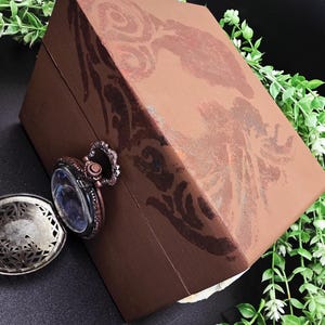 Puede incluir: Una caja de madera marrón con un diseño decorativo en la tapa. Un reloj de bolsillo de estilo vintage con una esfera transparente y detalles ornamentados está sujeto a la caja. Un óvalo decorativo plateado abierto está al lado de la caja.