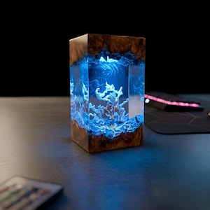 Peut inclure: Lampe rectangulaire en bois et résine avec un intérieur bleu brillant, ressemblant à un récif corallien. La lampe a un extérieur en grain de bois naturel et est éclairée par une lumière bleue, créant une pièce décorative unique.