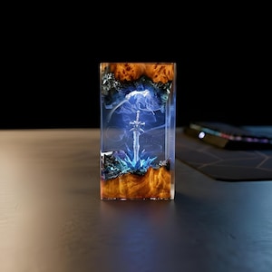 May include: A rectangular resin art piece featuring a miniature sword encased within. The artwork showcases a detailed sword design, set against a backdrop of blue and white smoky effects, with a base of textured wood and blue crystals.