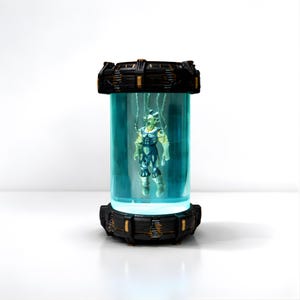 以下が含まれることがあります： 未来をテーマにしたディスプレイで、円筒形の透明な容器の中に吊り下げられたフィギュアが特徴です。フィギュアは緑色の肌色と青と白の衣装の人型です。容器は黒と金のアクセントが付いています。