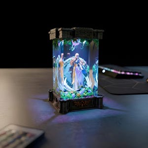 Puede incluir: Una vitrina iluminada y detallada con figuras de un juego de fantasía. La caja rectangular transparente tiene una base y una parte superior oscuras. La base tiene un emblema dorado. Las figuras están rodeadas de luces y follaje coloridos.