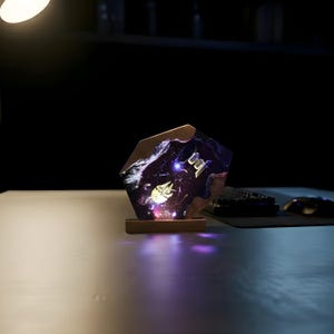 Peut inclure: Une œuvre d'art en résine illuminée unique, représentant une galaxie avec des vaisseaux spatiaux incrustés. L'œuvre est posée sur un socle en bois, avec un fond sombre et une source de lumière au-dessus. C'est un objet décoratif.