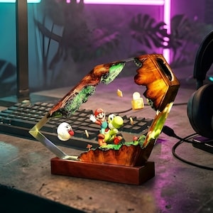 K&ouml;nnte beinhalten: Dekorative Lampe aus Holz und Kunstharz mit einer detaillierten Szene von Mario und Yoshi. Die Lampe hat einen rechteckigen Holzsockel und einen geometrischen Rahmen. Im Hintergrund befinden sich eine Tastatur und Kopfh&ouml;rer, mit Gaming-Thema.