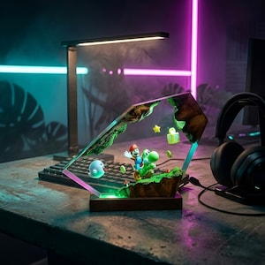 K&ouml;nnte beinhalten: Dekorative Lampe mit einer Szene mit Mario und Yoshi. Die Lampe hat einen Holzsockel und einen klaren, abgewinkelten Rahmen mit einer gr&uuml;nen und braunen Landschaft. Die Szene umfasst einen Geist, Sterne und schwebende Bl&ouml;cke. Eine schwarze Schreibtischlampe und Kopfh&ouml;rer sind ebenfalls sichtbar.