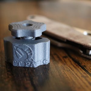 May include: A dark gray, 3D-printed plastic knob with a star-shaped top and a threaded base. The side of the knob features an embossed circular logo with a stylized helmet graphic and the text "T PAN" visible on the lower edge of the component.
