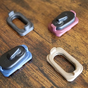 May include: Four rectangular mounting brackets with rounded corners. Two brackets are empty frames in black and beige. Two brackets are fitted with black oval-shaped locking mechanisms, one encased in a maroon base and one in a blue base.