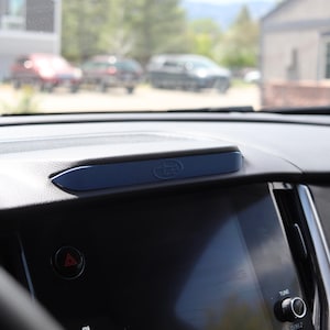 May include: A dark blue plastic dashboard trim piece with a subtle embossed logo, positioned centrally on the black textured dashboard of a vehicle, located directly above the integrated infotainment touchscreen display and air vents.