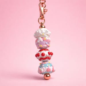 Puede incluir: Un dije decorativo con un cierre de mosquetón de metal en tono oro rosa y una hilera vertical de cuatro cuentas esféricas. Las cuentas presentan patrones coloridos en relieve en blanco, lavanda, rojo y azul claro con detalles dorados.