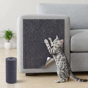 Puede incluir: Una lámina protectora adhesiva de color gris oscuro con rayas verticales aplicada en el lateral de un sofá gris claro, con un rollo a juego en primer plano. Un gato atigrado plateado está de pie sobre sus patas traseras junto al protector.