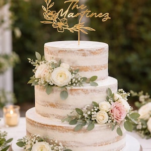 May include: A three-tiered naked cake with white frosting, decorated with white and light pink roses, baby's breath, and eucalyptus leaves. A gold-colored acrylic cake topper reads The Martinez in cursive script with a floral design.