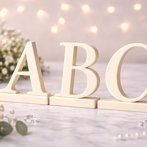 May include: Cream-colored wooden letters A, B, and C, each on a rectangular base. The letters are arranged in a row, with white flowers and a string of pearls in the background. Soft lighting creates a romantic ambiance.