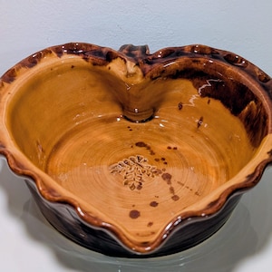 Puede incluir: Cuenco de cerámica en forma de corazón con borde ondulado. El cuenco tiene un interior marrón claro y un exterior marrón más oscuro. Un diseño floral decorativo está grabado en el centro. El borde tiene un borde texturizado marrón oscuro.