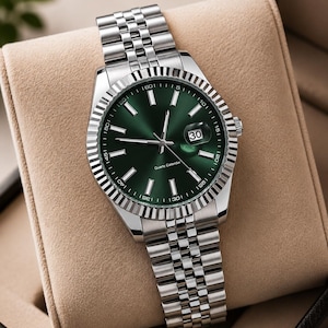 May include: A silver wristwatch with a green dial and a date display at the 3 o'clock position. The watch has a fluted bezel and a stainless steel bracelet. The watch is displayed in a tan watch box.