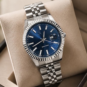 May include: A stainless steel wristwatch with a blue dial and a fluted bezel. The watch features a date display at the 3 o'clock position and a silver-tone bracelet. The watch is presented in a beige box.