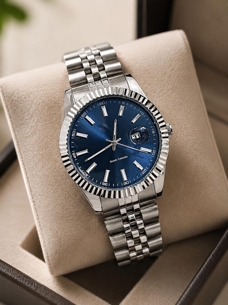 May include: A stainless steel wristwatch with a blue dial and a fluted bezel. The watch features a date display at the 3 o'clock position and a silver-tone bracelet. The watch is presented in a beige box.