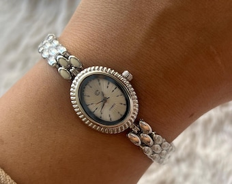 May include: A silver-tone quartz wristwatch featuring an oval-shaped face with a sunburst dial, a fluted bezel, and a link bracelet composed of small, rounded, metallic segments. The watch dial displays the letter C and the word QUARTZ.