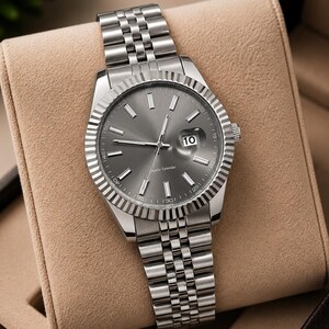 May include: A silver-toned wristwatch with a gray face and a date display at the 3 o'clock position. The watch has a fluted bezel and a stainless steel band. The watch is displayed in a tan watch box.
