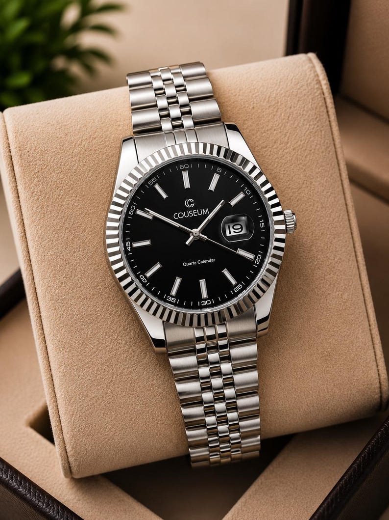 May include: A silver-toned wristwatch with a black face and a date display at the 3 o'clock position. The watch features a fluted bezel and a stainless steel band. The watch is displayed in a tan watch box.