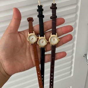 May include: Three small wristwatches with gold-colored cases and white faces. The watch bands are brown, black, and dark brown. The watches are displayed against a neutral background.