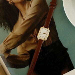May include: A gold-toned rectangular watch with a white face and brown leather band. The watch has a classic design with a gold buckle. The watch is displayed on a magazine with a woman in the background.