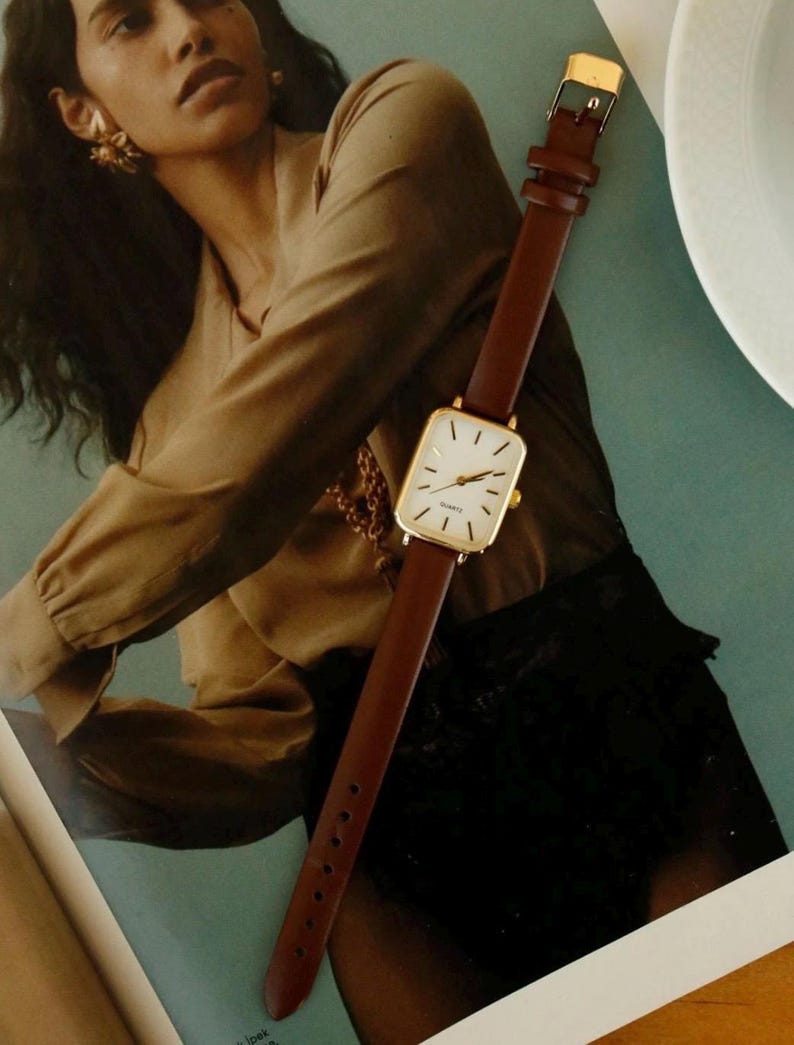 May include: A gold-toned rectangular watch with a white face and brown leather band. The watch has a classic design with a gold buckle. The watch is displayed on a magazine with a woman in the background.