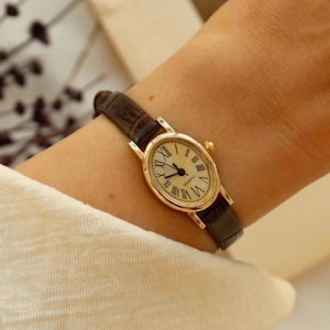 May include: A gold-toned wristwatch with a brown leather band. The watch face is oval-shaped with Roman numeral hour markers and black hands. The watch is worn on a wrist, with a cream-colored fabric in the background.