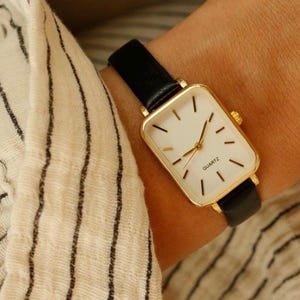 May include: A gold-toned rectangular quartz watch with a white face and black hands and markers. The watch has a black leather band. The watch is on a wrist, and the background is a striped fabric.