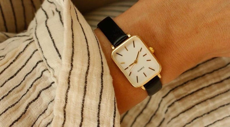 May include: A gold-toned rectangular quartz watch with a white face and black hands and markers. The watch has a black leather band. The watch is on a wrist, and the background is a striped fabric.