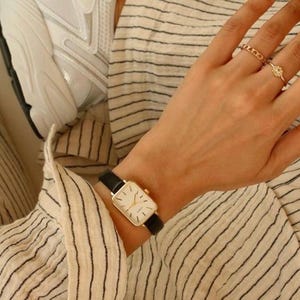 May include: A gold rectangular watch with a black leather band is worn on a wrist. The watch face is white with black hands and markers. The person is wearing rings on their fingers. The person is wearing a striped shirt.