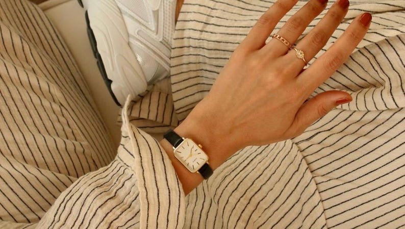 May include: A gold rectangular watch with a black leather band is worn on a wrist. The watch face is white with black hands and markers. The person is wearing rings on their fingers. The person is wearing a striped shirt.