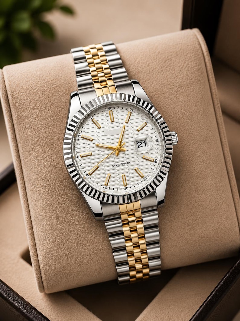 May include: A silver and gold wristwatch with a white dial and a date display at the 3 o'clock position. The watch has a fluted bezel and a two-tone stainless steel and gold-tone link bracelet. The watch is displayed in a tan watch box.