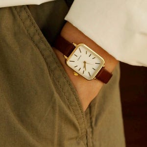May include: A gold-toned rectangular quartz watch with a white face and black hour markers. The watch has a brown leather band. The watch is being worn on a wrist, with a white shirt and olive green pants in the background.