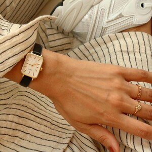 May include: A gold rectangular wristwatch with a black leather band. The watch has a white face with gold hands and markers. The person is wearing gold rings. The background includes a striped shirt and a white sneaker.