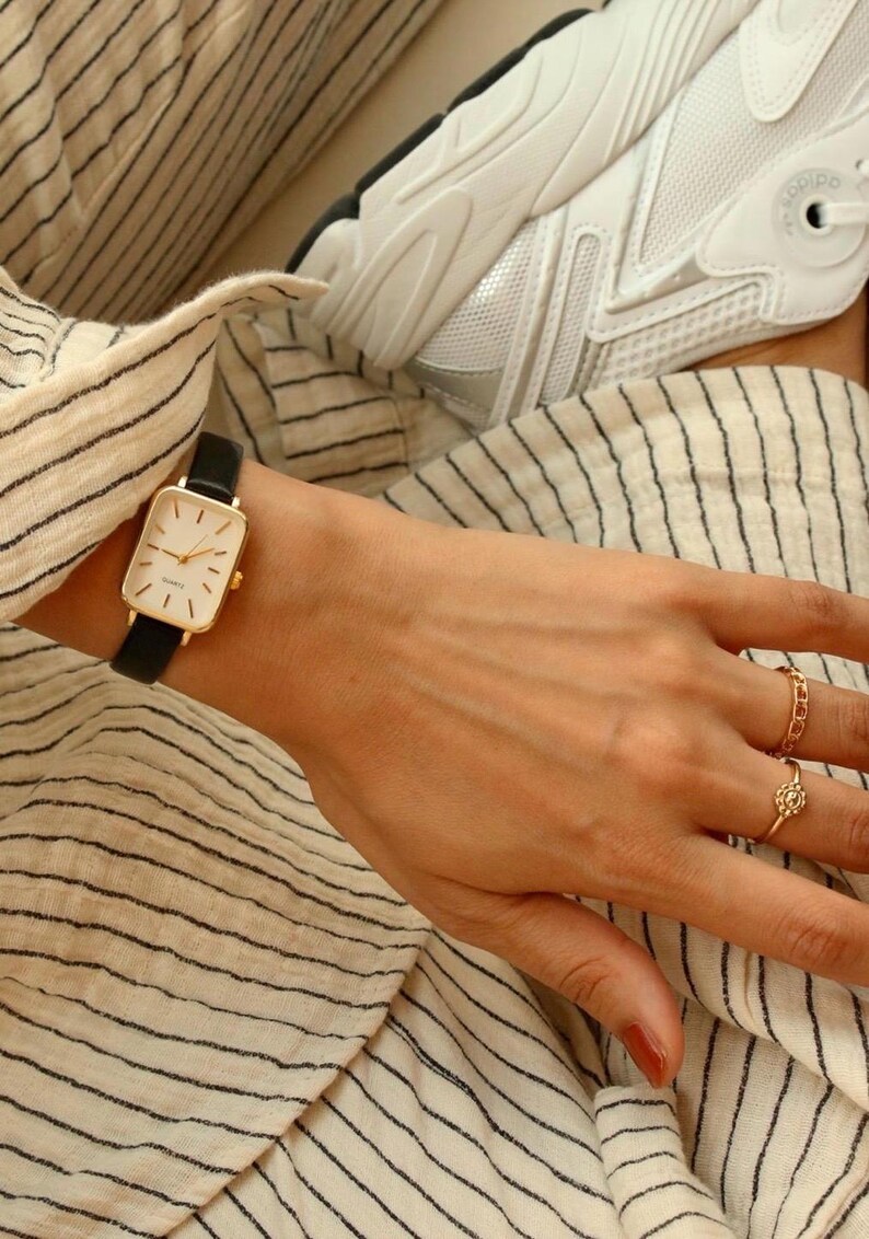 May include: A gold rectangular wristwatch with a black leather band. The watch has a white face with gold hands and markers. The person is wearing gold rings. The background includes a striped shirt and a white sneaker.