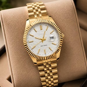 May include: Gold-toned wristwatch with a white dial and a date display at the 3 o'clock position. The watch features a fluted bezel and a gold link bracelet. The watch is presented in a tan watch box.