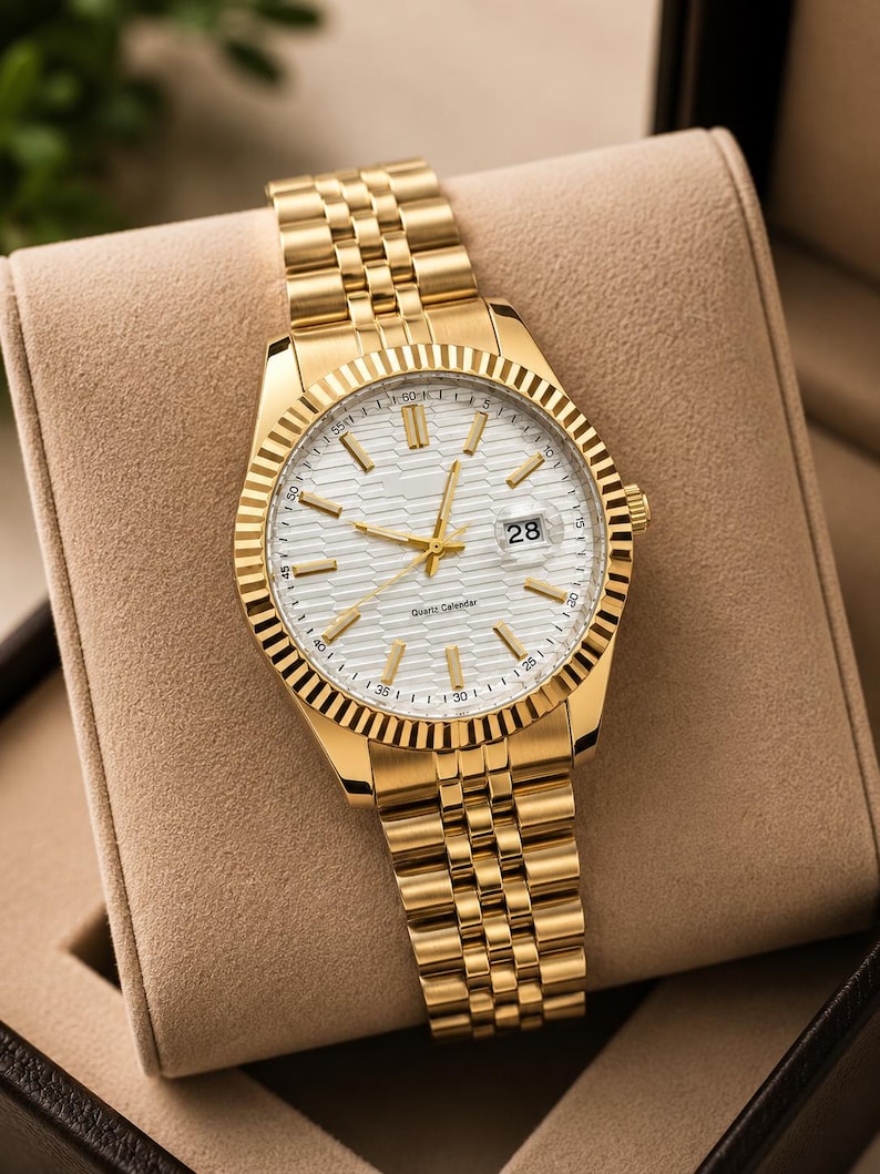 May include: Gold-toned wristwatch with a white dial and a date display at the 3 o'clock position. The watch features a fluted bezel and a gold link bracelet. The watch is presented in a tan watch box.
