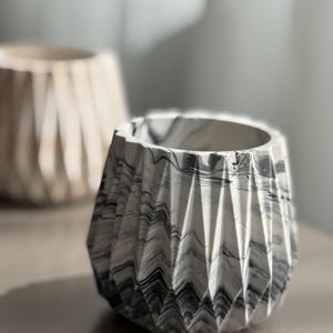 May include: A close-up of two decorative geometric planters. The front planter has a marbled black and white design. The second planter is out of focus and has a light beige color. Both planters have a faceted design.