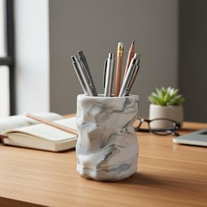 May include: A white and gray marble-patterned pen holder filled with pens and pencils sits on a wooden desk. A laptop, open notebook, and small potted plant are also on the desk, suggesting a workspace.